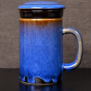 Könnte beinhalten: Eine blaue Keramik-Tasse mit passendem Deckel. Die Tasse hat einen Henkel und einen Farbverlauf, der von Braun zu Blau übergeht. Die Tasse ist für Heißgetränke konzipiert.