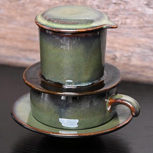 Könnte beinhalten: Ein Keramik-Kaffeefilter-Set in Grün- und Brauntönen. Das Set besteht aus einem Deckel, einem Filter, einer Untertasse und einer Tasse mit Henkel. Die Teile sind gestapelt, was das Design und die Funktionalität des Kaffeezubereitungssets zeigt.