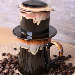 May include: A dark brown ceramic coffee filter and mug set with a dripped glaze effect. The set includes a lid, filter, and mug, surrounded by coffee beans. The mug has a curved handle.