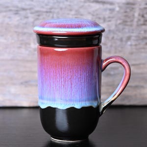 May include: A ceramic tea mug with a lid, featuring a gradient of colors from red to purple to blue, with a black base. The mug has a handle and is designed for brewing tea.