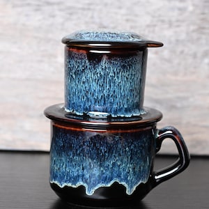 Puede incluir: Un juego de filtro de café de cerámica con un esmalte azul oscuro y un efecto de goteo. El juego incluye una taza con asa, un filtro y una tapa. El esmalte hace una transición de marrón oscuro a azul.