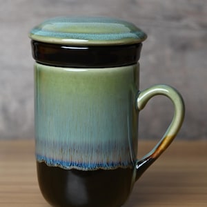 May include: A ceramic tea mug with a lid, featuring a reactive glaze that transitions from a dark brown base to a light green and blue upper section. The mug includes a curved handle and a matching lid with a smooth, glossy finish throughout.