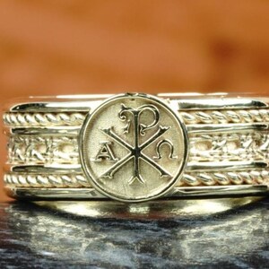 May include: A gold ring with a detailed design. The ring features a central circular emblem with religious symbols, surrounded by intricate rope and patterned bands. The ring is displayed against a blurred brown background.