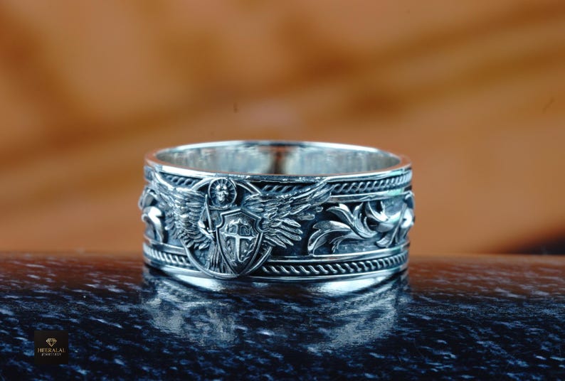 May include: A detailed silver ring with an embossed design. The ring features an angel with outstretched wings, a shield, and decorative scrollwork. The ring is set against a blurred background.