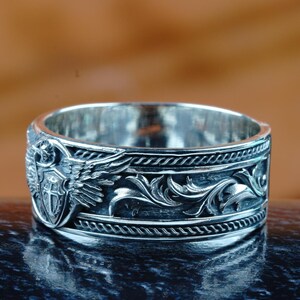 May include: Silver ring with an engraved design. The ring features an emblem with wings and a shield, along with a detailed floral pattern. The ring has a polished finish and is displayed on a reflective surface.