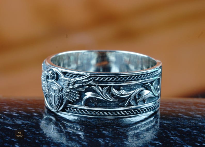 May include: Silver ring with an engraved design. The ring features an emblem with wings and a shield, along with a detailed floral pattern. The ring has a polished finish and is displayed on a reflective surface.