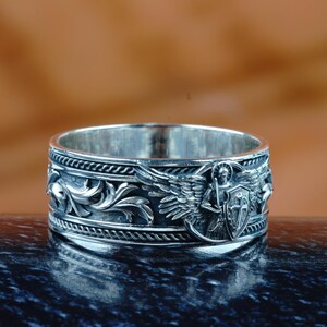May include: A silver ring with detailed relief carvings. The ring features an angel with wings, a shield, and ornate floral patterns. The ring is displayed on a dark, reflective surface against a blurred brown background.