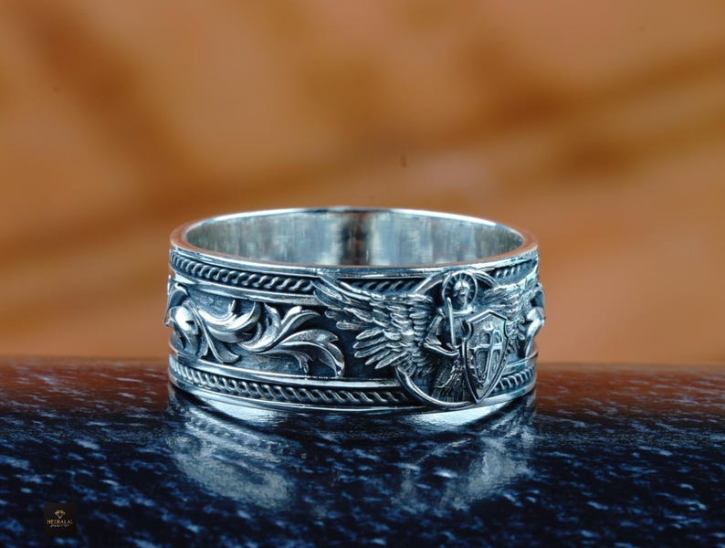 May include: A silver ring with detailed relief carvings. The ring features an angel with wings, a shield, and ornate floral patterns. The ring is displayed on a dark, reflective surface against a blurred brown background.