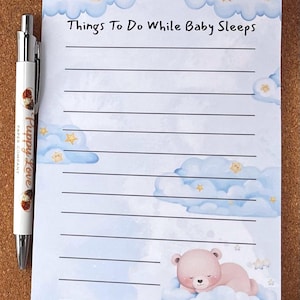 May include: A notepad with the text "Things To Do While Baby Sleeps" features a watercolor design of clouds, stars, and a sleeping teddy bear. A Puppy Love pen rests on the side. The notepad has lined pages for writing.