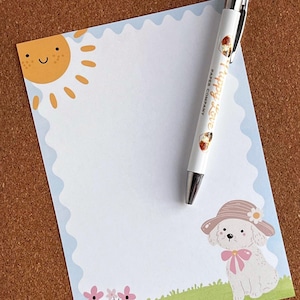 Puede incluir: Un juego de papelería con un bloc de notas y un bolígrafo. El bloc de notas tiene un borde festoneado y un sol de dibujos animados, un perro y flores. El bolígrafo es blanco con las palabras "Happy Love Paper Company" y un diseño de postre.