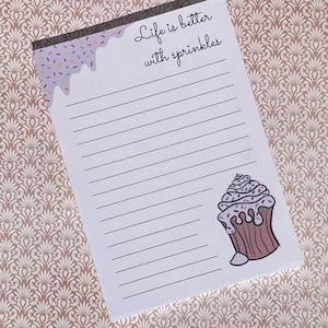 May include: A lined notepad with the text "Life is better with sprinkles." The top border has a purple design with colorful sprinkles. A cupcake illustration is on the bottom right corner.