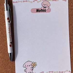 May include: A white notepad with a pink and yellow bone border and a pink oval that says "Notes". A pen with the text "Puppy Love Paper Company" is next to the notepad. The notepad has a cartoon puppy illustration.