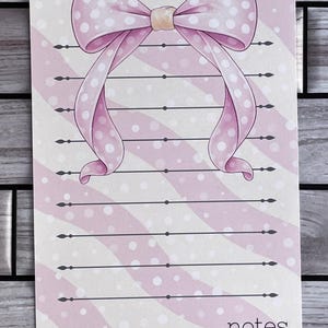 Può includere: Un blocco note a righe rosa e bianche con un grande fiocco rosa con pois bianchi. Il blocco note ha linee orizzontali per la scrittura e la parola "notes" nell'angolo in basso a destra.