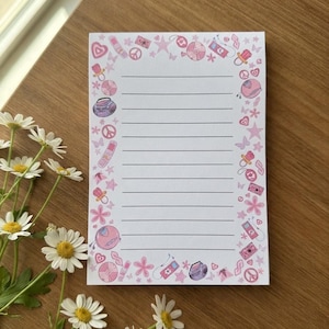 May include: A rectangular notepad with horizontal black lines and a decorative border featuring various pink and purple illustrations, including peace signs, stars, hearts, butterflies, cassette players, and pacifiers on a white background.