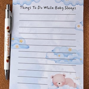 May include: A notepad with the text "Things To Do While Baby Sleeps" in black font. The notepad features a light blue watercolour design with clouds, stars, and a sleeping bear illustration. A pen with the text "Puppy Love Paper Company" is on the left.