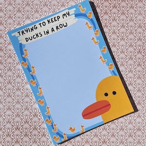 Könnte beinhalten: Ein Notizblock mit blauem Hintergrund und dem Text "TRYING TO KEEP MY DUCKS IN A ROW". Der Notizblock ist mit gelben Entenillustrationen und einem großen gelben Entenkopf verziert. Der Notizblock liegt auf einer gemusterten Oberfläche.
