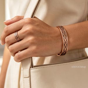 Könnte beinhalten: Ein kupferfarbener Metall-Armreif und ein passender Ring, beide mit einem geprägten, sich wiederholenden Unendlichkeitsmuster und abgedunkelten, vertieften Details. Der Text AMNATshop ist in der unteren rechten Ecke des Bildes zu sehen.