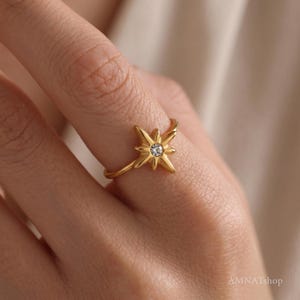May include: A gold-toned ring featuring a central starburst motif with a small, clear, round-cut gemstone at its center. The starburst design has eight points of varying lengths, and the band is thin and smooth, encircling the finger.