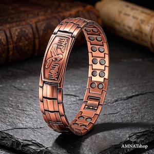 Op de afbeelding: Een koperkleurige metalen schakelarmband met een rechthoekige centrale plaat voorzien van een reliëf van een adelaar en een bos. De armband is opgebouwd uit onderling verbonden rechthoekige segmenten met zichtbare ronde magnetische inzetstukken.