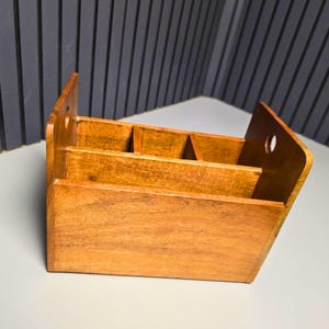 May include: A medium-brown wooden desktop organizer featuring a rectangular base, three internal compartments for storage, and two tall side panels, each with a circular cutout handle near the top edge for easy carrying and transport.