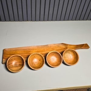 Peut inclure: Une longue planche de service rectangulaire en bois avec une poignée à une extrémité, présentant un grain de bois brun naturel. Quatre petits bols ronds en bois aux tons bruns assortis sont disposés en rangée le long de la planche.