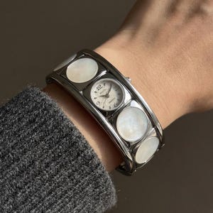 Könnte beinhalten: Eine silberfarbene Armbanduhr mit einem kleinen runden Zifferblatt und weißen Perlmuttakzenten. Das Zifferblatt zeigt die Zahl 12 und den Markennamen "Wocti". Die Uhr wird am Handgelenk getragen.