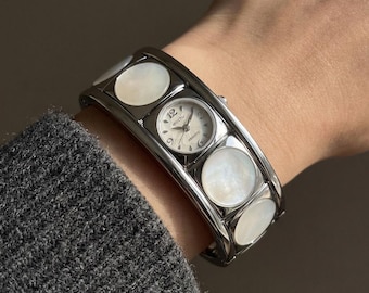 May include: A silver-toned cuff watch with a small round watch face and white mother-of-pearl accents. The watch face displays the number 12 and the brand name "Wocti". The watch is worn on a wrist.