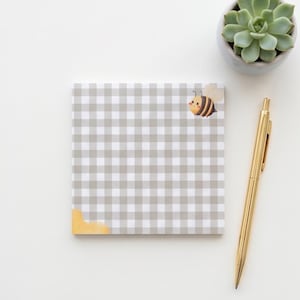 May include: A square notepad featuring a gray and white gingham pattern, a small cartoon bee illustration in the top right corner, and a yellow watercolor-style accent in the bottom left corner, accompanied by a gold-toned metal ballpoint pen.