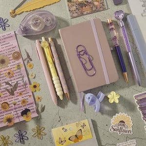 Puede incluir: Un surtido de artículos de papelería en colores pastel, que incluyen bolígrafos, un cuaderno, pegatinas y un dispensador de cinta adhesiva. Los artículos presentan motivos florales y de mariposas, creando una estética caprichosa.