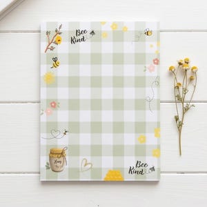 May include: A notepad with a green and white gingham pattern, decorated with illustrations of bees, flowers, and a honey jar. The words "Bee Kind" are written on the top and bottom of the notepad.
