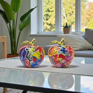 Puede incluir: Dos esculturas decorativas de manzana con tallos dorados. Una manzana presenta un diseño de estilo grafiti con colores vibrantes, mientras que la otra muestra un patrón floral en rojo, amarillo y azul. Están sobre una mesa de cristal.