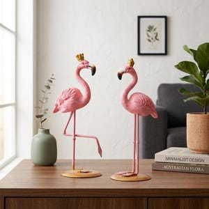 May include: Two pink flamingo figurines standing on circular gold bases. Each flamingo features a textured body, a long neck, and a small gold crown perched on its head. One flamingo stands on one leg, while the other stands on two legs.