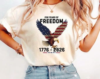 May include: A cream-colored short-sleeve t-shirt featuring a graphic of a bald eagle with wings patterned like the American flag. Above the eagle, the text reads 250 YEARS OF FREEDOM, and below the eagle, the text reads 1776 - 2026 with stars.