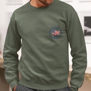 May include: An olive green crewneck sweatshirt featuring a small circular graphic on the left chest. The design includes a map of the United States filled with the American flag pattern, surrounded by a ring of blue stars with the text 1776 and 2026.