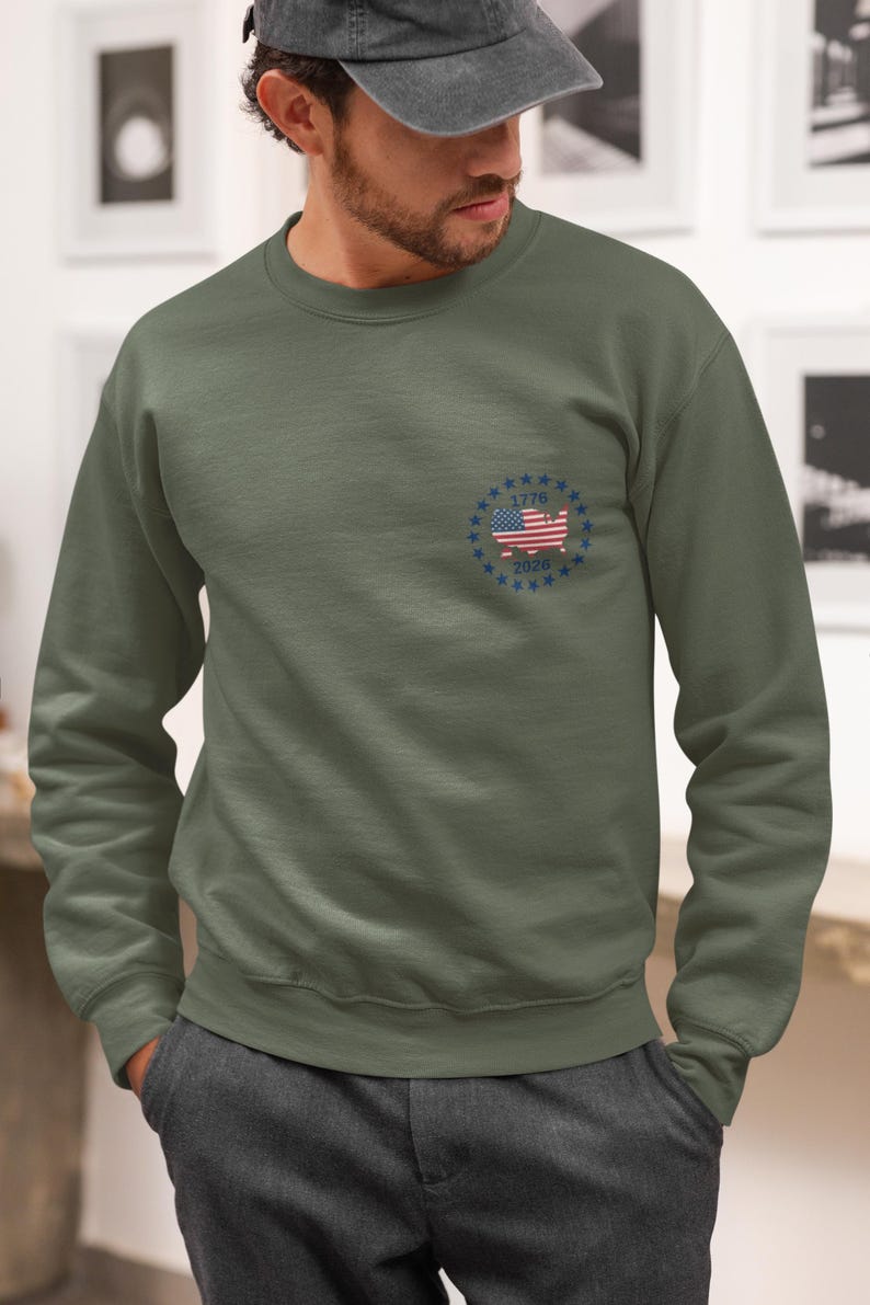 May include: An olive green crewneck sweatshirt featuring a small circular graphic on the left chest. The design includes a map of the United States filled with the American flag pattern, surrounded by a ring of blue stars with the text 1776 and 2026.