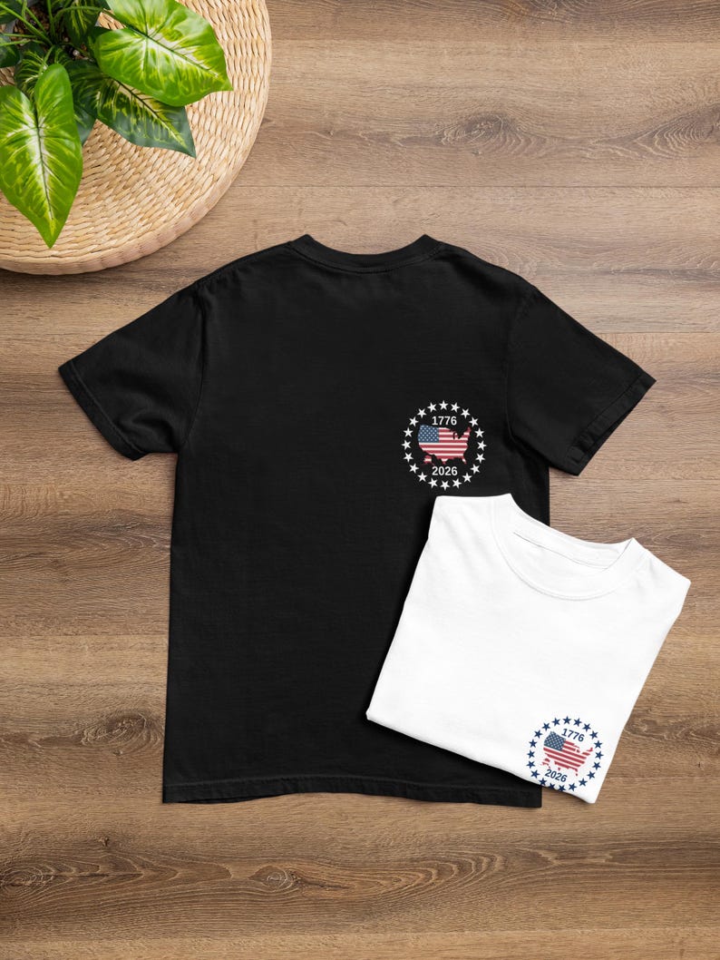 May include: Two t-shirts, one black and one white, are displayed on a wooden surface. Each shirt features a small graphic of the American flag within a circle of stars, with the numbers "1776" and "2026" below.