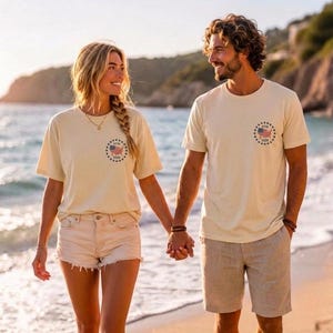 May include: Two cream-colored short-sleeve t-shirts featuring a circular graphic on the left chest. The graphic displays a small American flag inside a ring of stars with the text 1776 above and 2026 below the flag. The shirts are made of cotton.