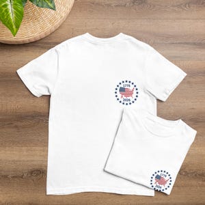 May include: A white short-sleeved t-shirt featuring a circular graphic on the left chest. The design includes a map of the United States filled with the American flag pattern, surrounded by a ring of blue stars, with the text 1776 above and 2026 below.