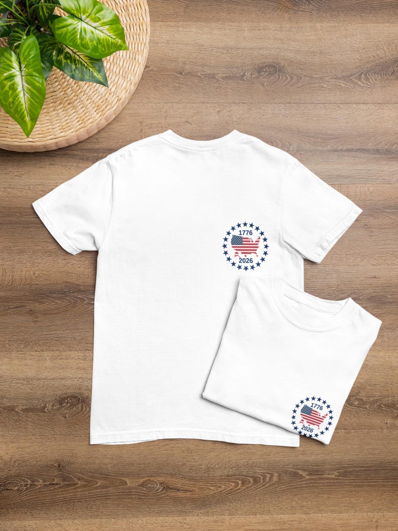 May include: A white short-sleeved t-shirt featuring a circular graphic on the left chest. The design includes a map of the United States filled with the American flag pattern, surrounded by a ring of blue stars, with the text 1776 above and 2026 below.