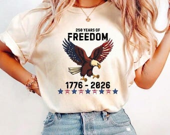 May include: A cream-colored t-shirt featuring a graphic of a bald eagle with wings spread. Above the eagle, the text reads 250 YEARS OF FREEDOM. Below the eagle, the text reads 1776-2026, followed by a row of seven small stars in red, white, and blue.