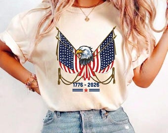 May include: A cream-colored t-shirt featuring a graphic of a bald eagle centered between two crossed American flags. Below the eagle, the text 1776-2026 is printed in blue, separated by a small red star and two horizontal blue lines.