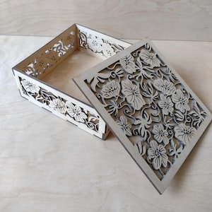 Könnte beinhalten: Eine hellfarbene Holzkiste mit einem floralen Design, das in den Deckel und die Seiten geschnitten ist. Der Deckel ist teilweise geöffnet und gibt den Innenraum frei. Das florale Muster ist aufwendig und detailliert.