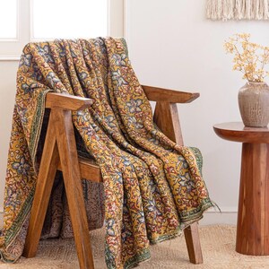 May include: A wooden chair with a yellow floral blanket. The blanket has a repeating pattern of blue, red, and white flowers on a yellow backdrop. The chair is made of light brown wood and rests on a woven rug. The blanket's edge has a green border.