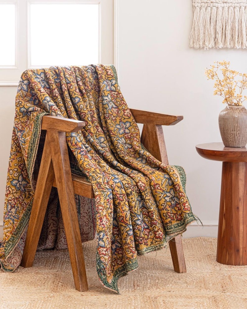 May include: A wooden chair with a yellow floral blanket. The blanket has a repeating pattern of blue, red, and white flowers on a yellow backdrop. The chair is made of light brown wood and rests on a woven rug. The blanket's edge has a green border.