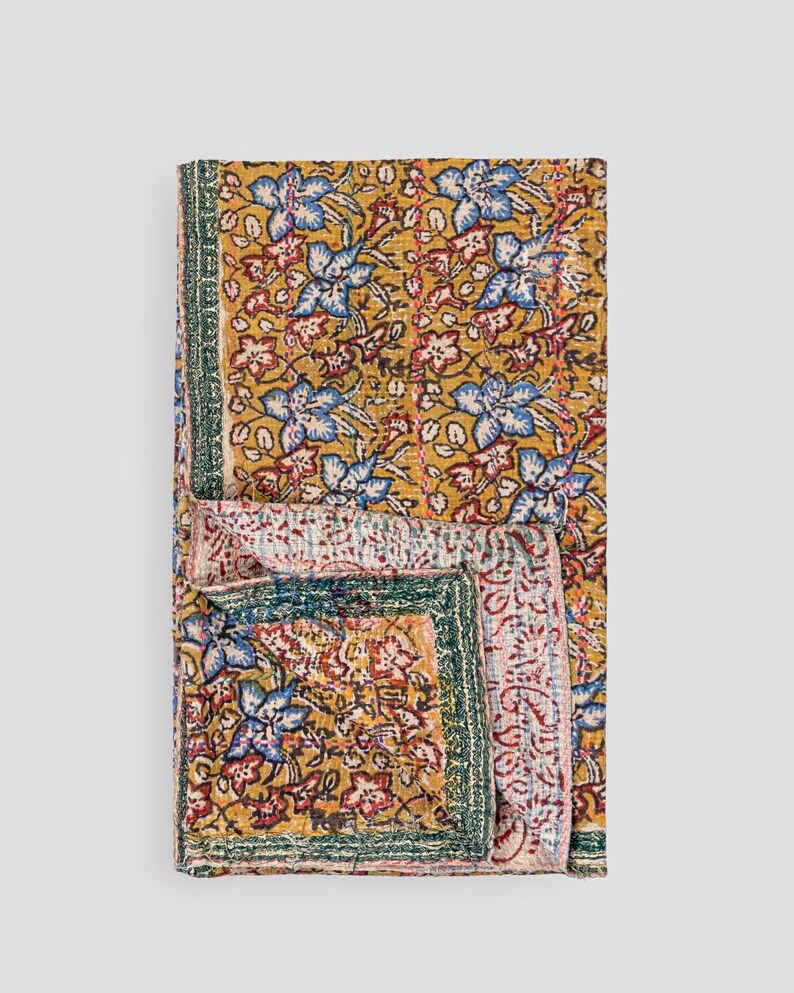 May include: A folded quilt with a floral pattern in gold, blue, red, and green. The quilt displays a repeating design of stylized flowers and leaves. It has a green border. The quilt is folded to show the pattern.