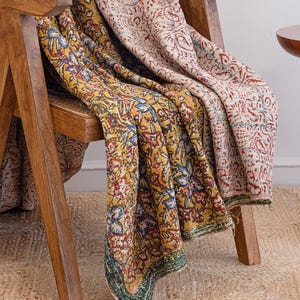 May include: A wooden chair with two patterned blankets draped over it. One blanket is yellow with blue, red, and green floral designs. The other is beige with red floral patterns. The blankets are folded over the chair.