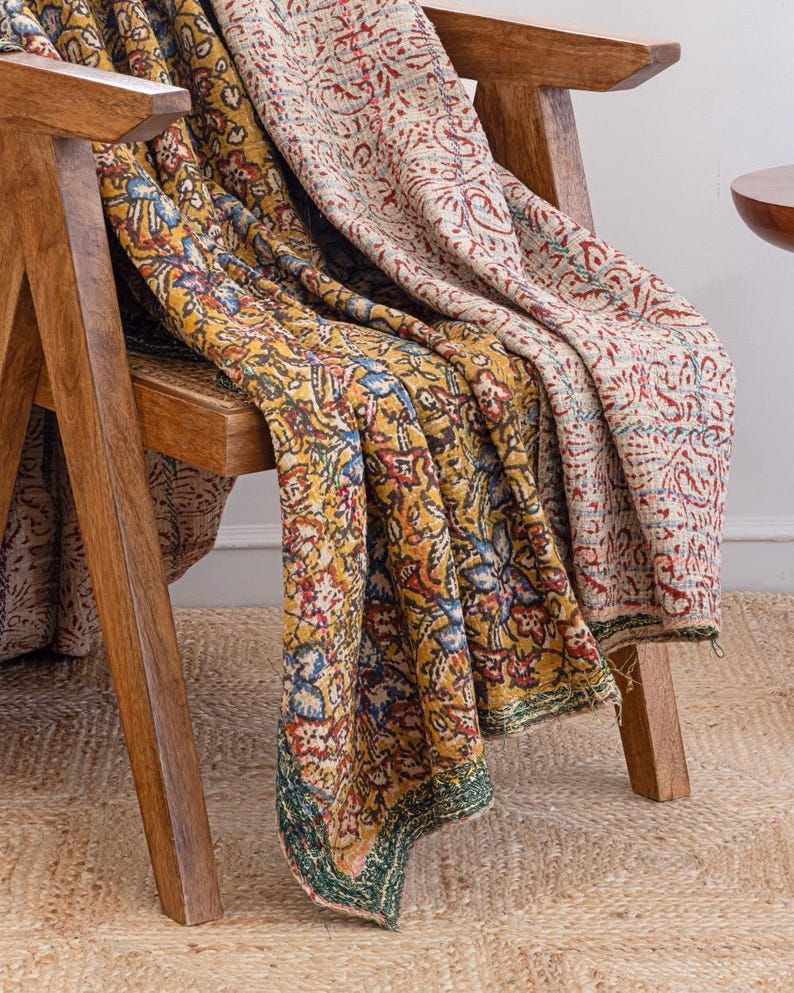 May include: A wooden chair with two patterned blankets draped over it. One blanket is yellow with blue, red, and green floral designs. The other is beige with red floral patterns. The blankets are folded over the chair.