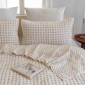 Puede incluir: Juego de cama color crema con un patrón de puntada de cruz en negro. El conjunto incluye una colcha y almohadas. Un libro titulado "Home Therapy" descansa sobre la cama. El ambiente es neutro.