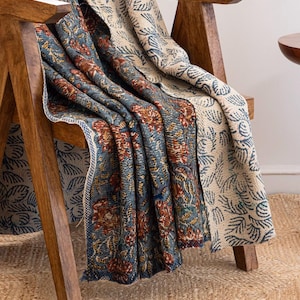 May include: A reversible throw blanket draped over a wooden chair. One side features a blue floral pattern with red and yellow accents, while the other side has a cream-coloured design with blue leaf motifs. The blanket is made of a soft, textured fabric.