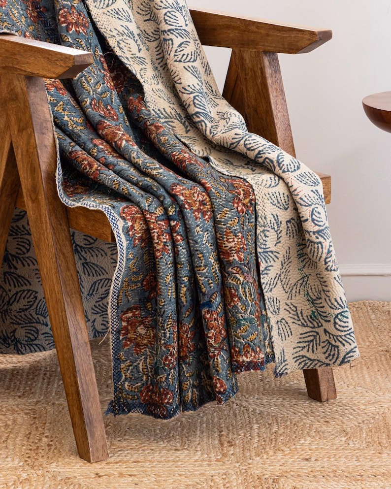 May include: A reversible throw blanket draped over a wooden chair. One side features a blue floral pattern with red and yellow accents, while the other side has a cream-coloured design with blue leaf motifs. The blanket is made of a soft, textured fabric.